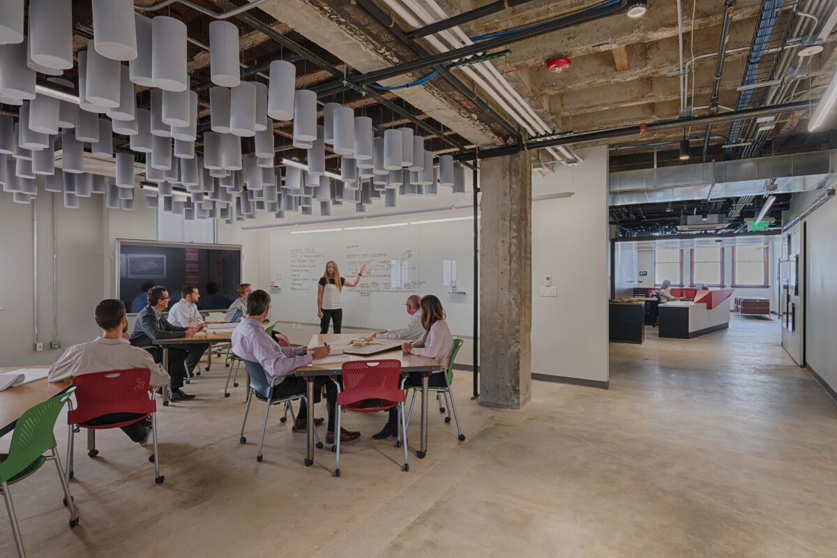 employees seated at tables during a meeting while a presenter speaks at a whiteboard in an open industrial-style office