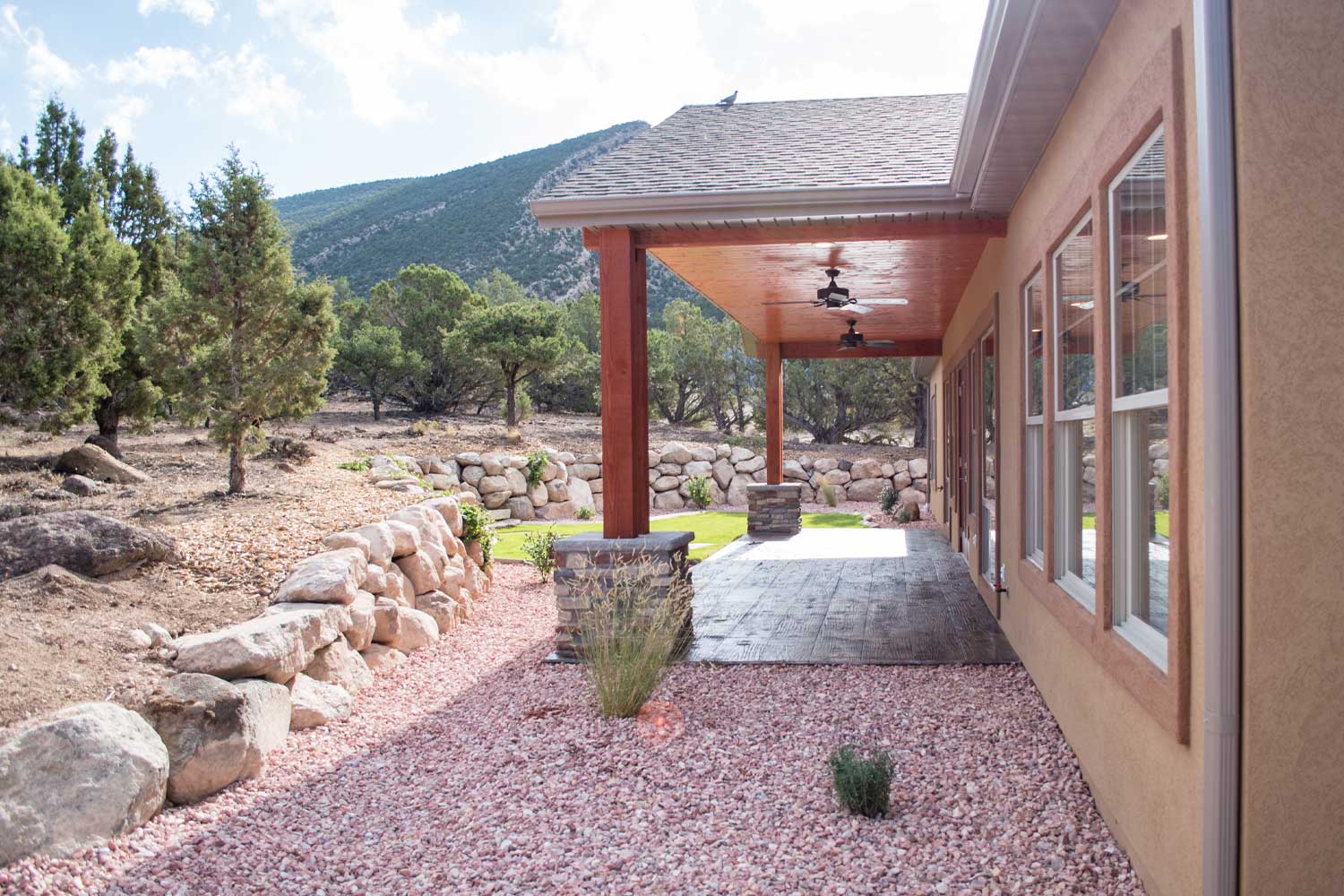 back patio exterior, small red rock gravel, smooth stamped dark brown concrete, rock retaining wall and pine trees, blue sky with light clouds