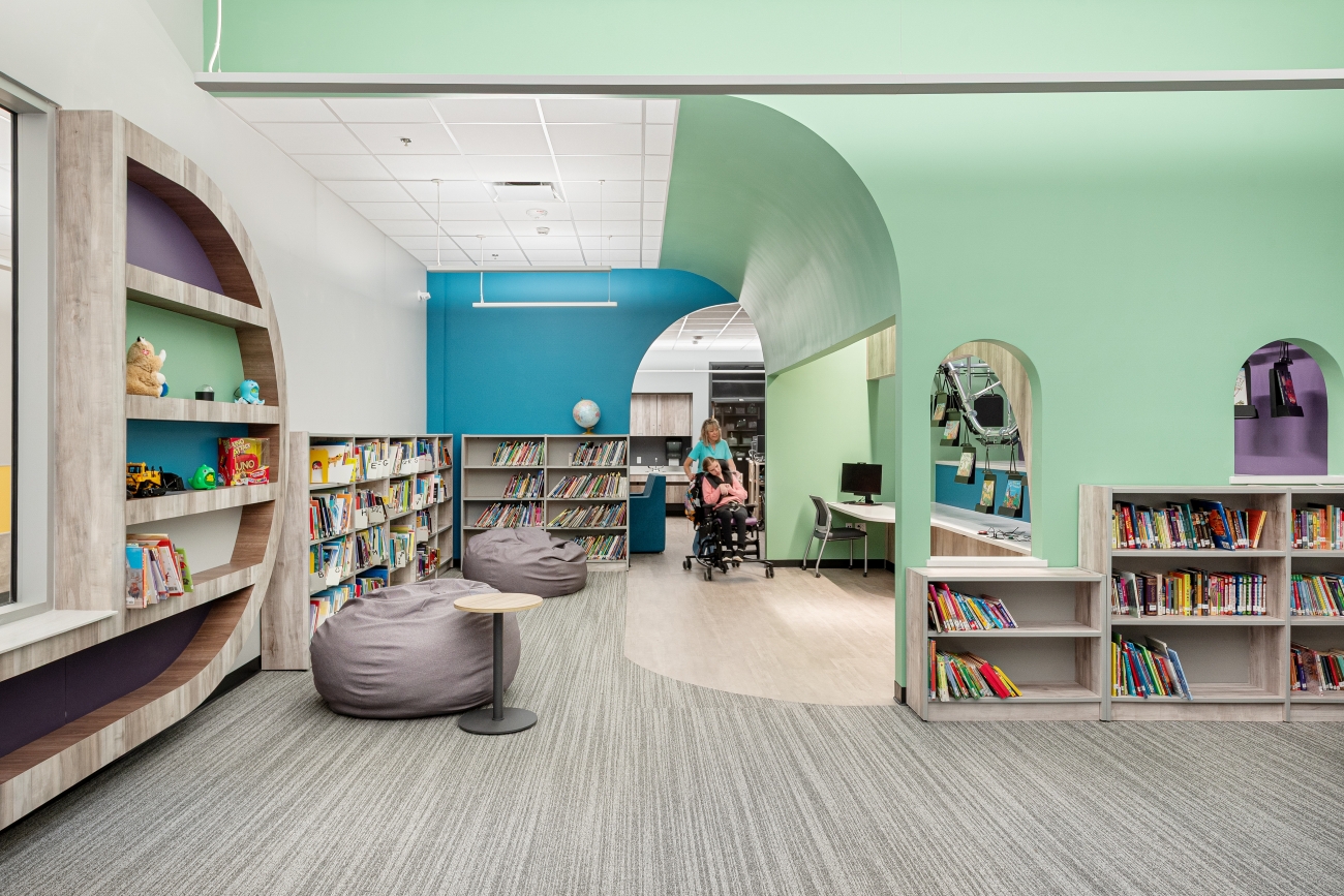 Library service desk with an overhead book conveyor system, integrated shelving, curved millwork counter, and adjacent reading and circulation areas.