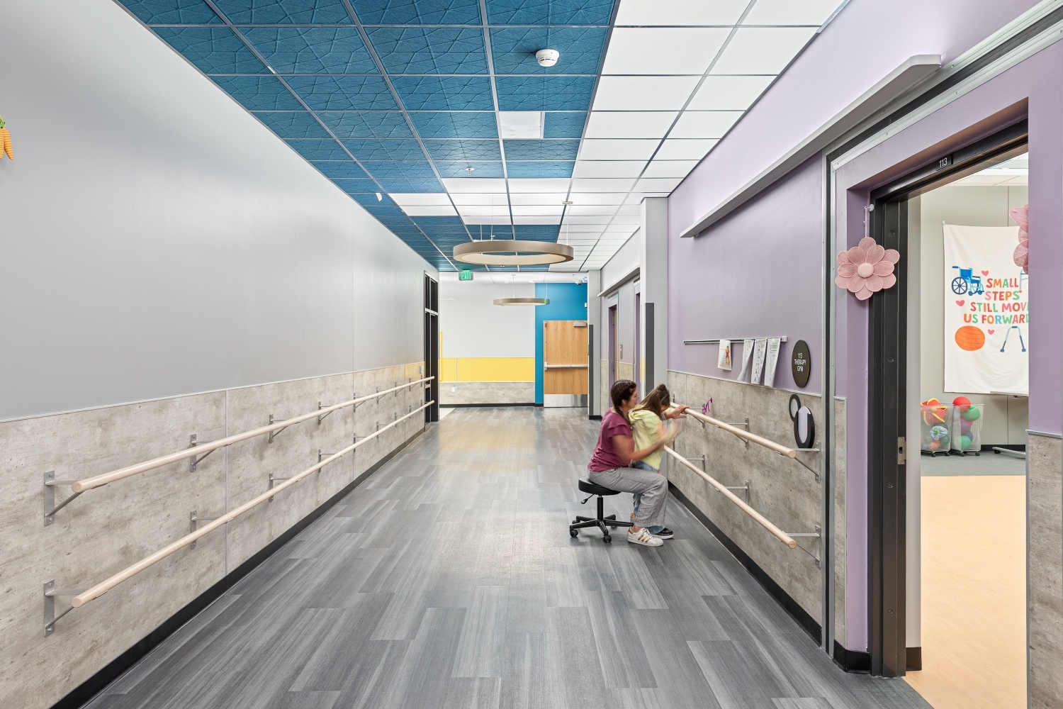 Therapy corridor with dual handrails along the walls, resilient flooring, acoustic ceiling panels, circular suspended light fixtures, and adjacent classroom doorways.