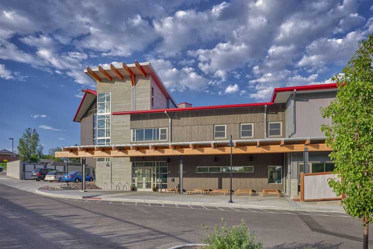 Exterior view of the Great Work Montessori PK-5 School building with a modern architectural design featuring large windows, a wooden canopy, and red roof accents under a partly cloudy sky.