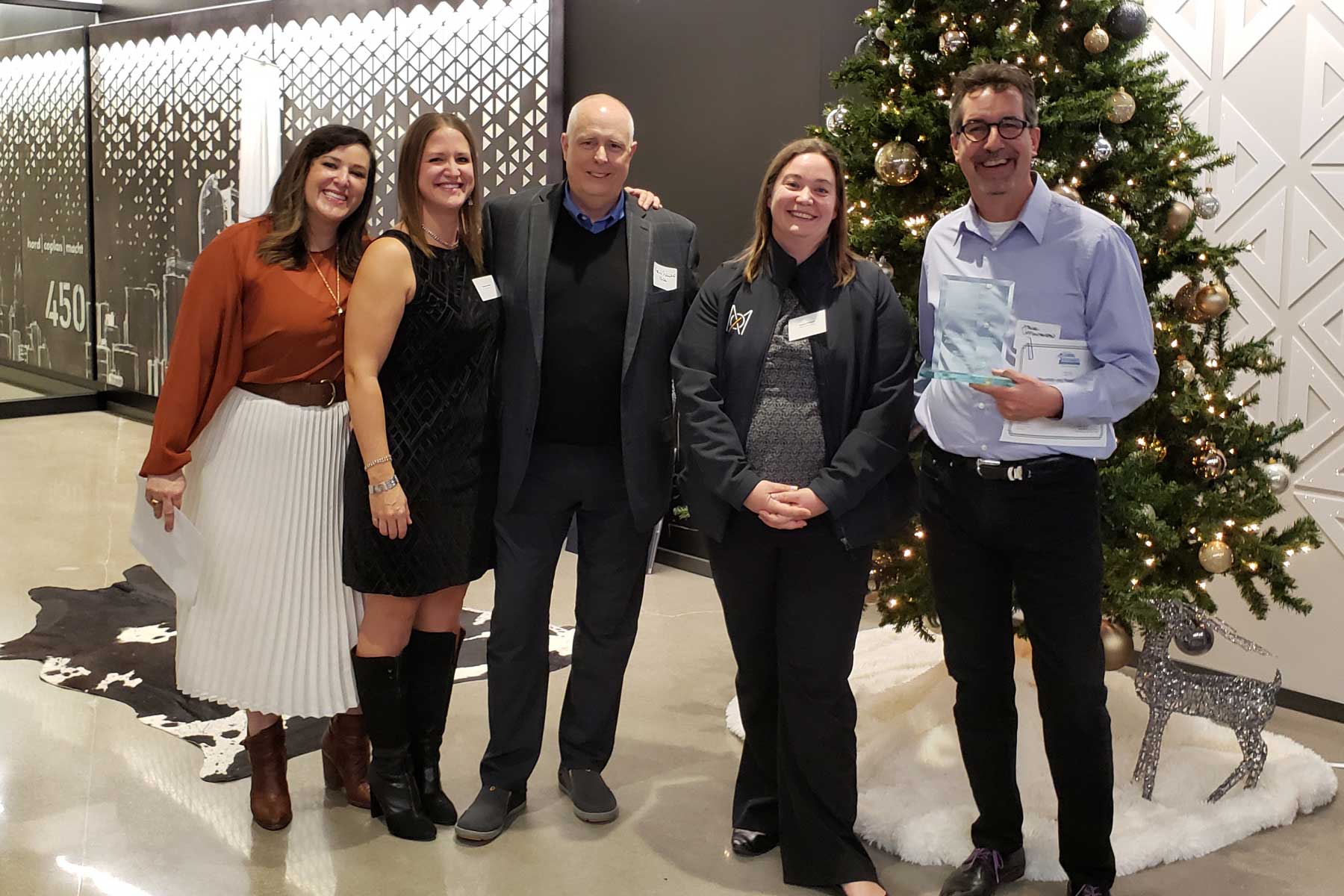 Five people standing in a lobby area in front of a decorated Christmas tree, smiling and posing for a photo, and one person on the right is holding an award.