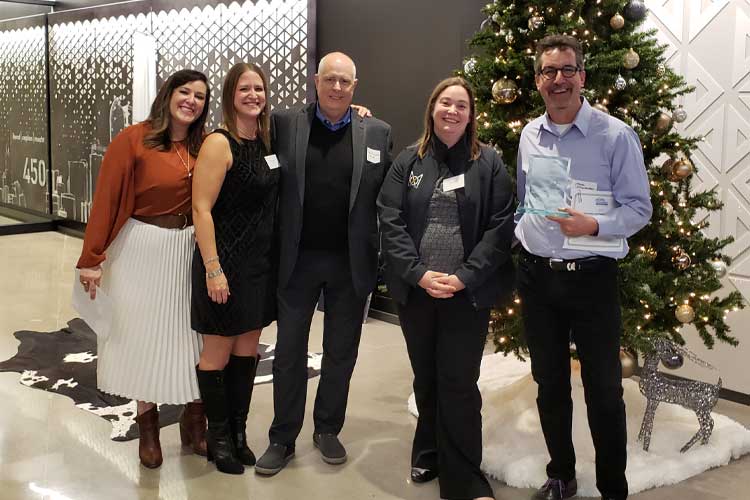 Five people standing in a lobby area in front of a decorated Christmas tree, smiling and posing for a photo, and one person on the right is holding an award.