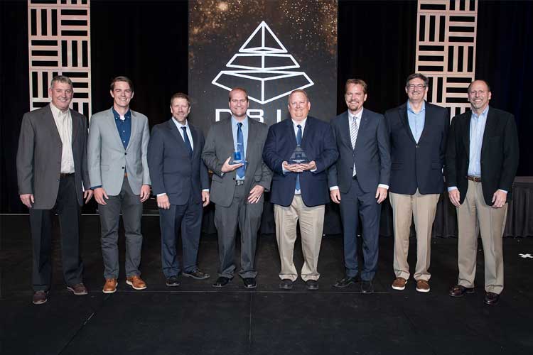 A group of men in business attire standing in a row on a stage at a DBIA award ceremony holding awards, with a DBIA logo displayed on a screen behind them.