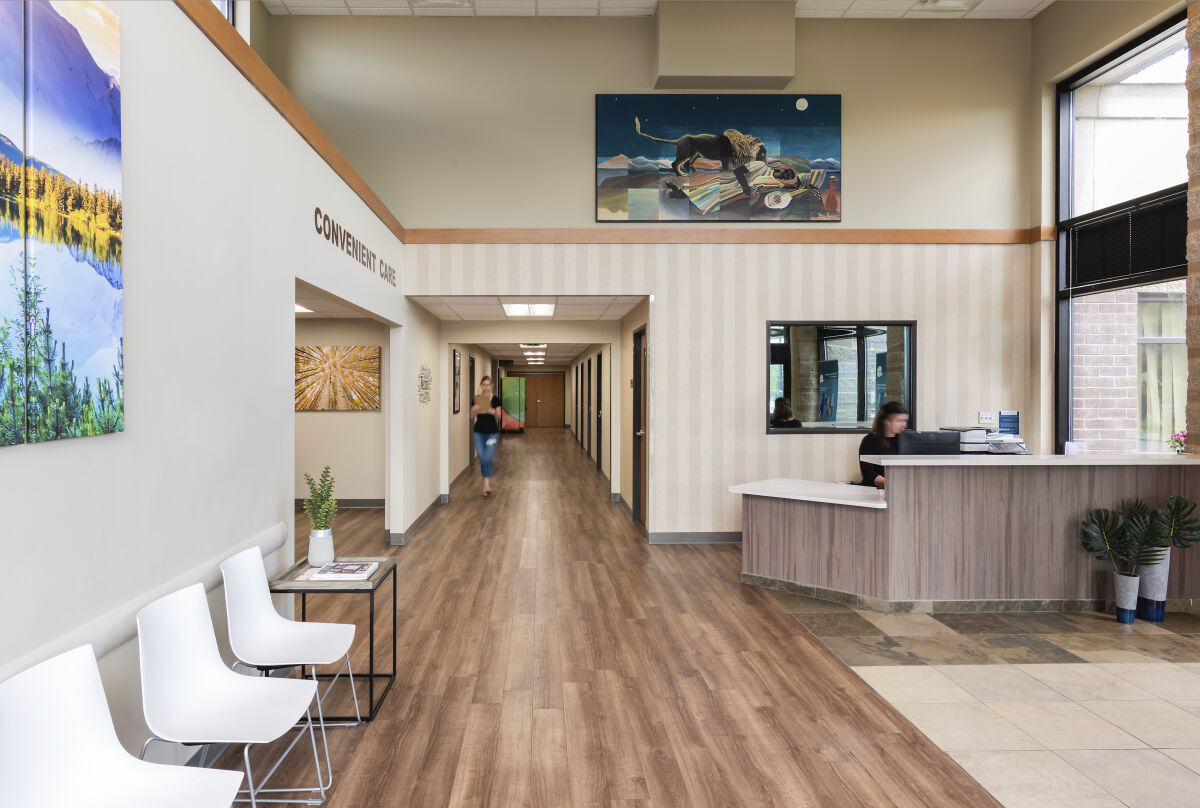 Lobby interior with a wood-look floor, reception desk with a glass window, white waiting chairs, framed artwork, and a central corridor leading to exam rooms.