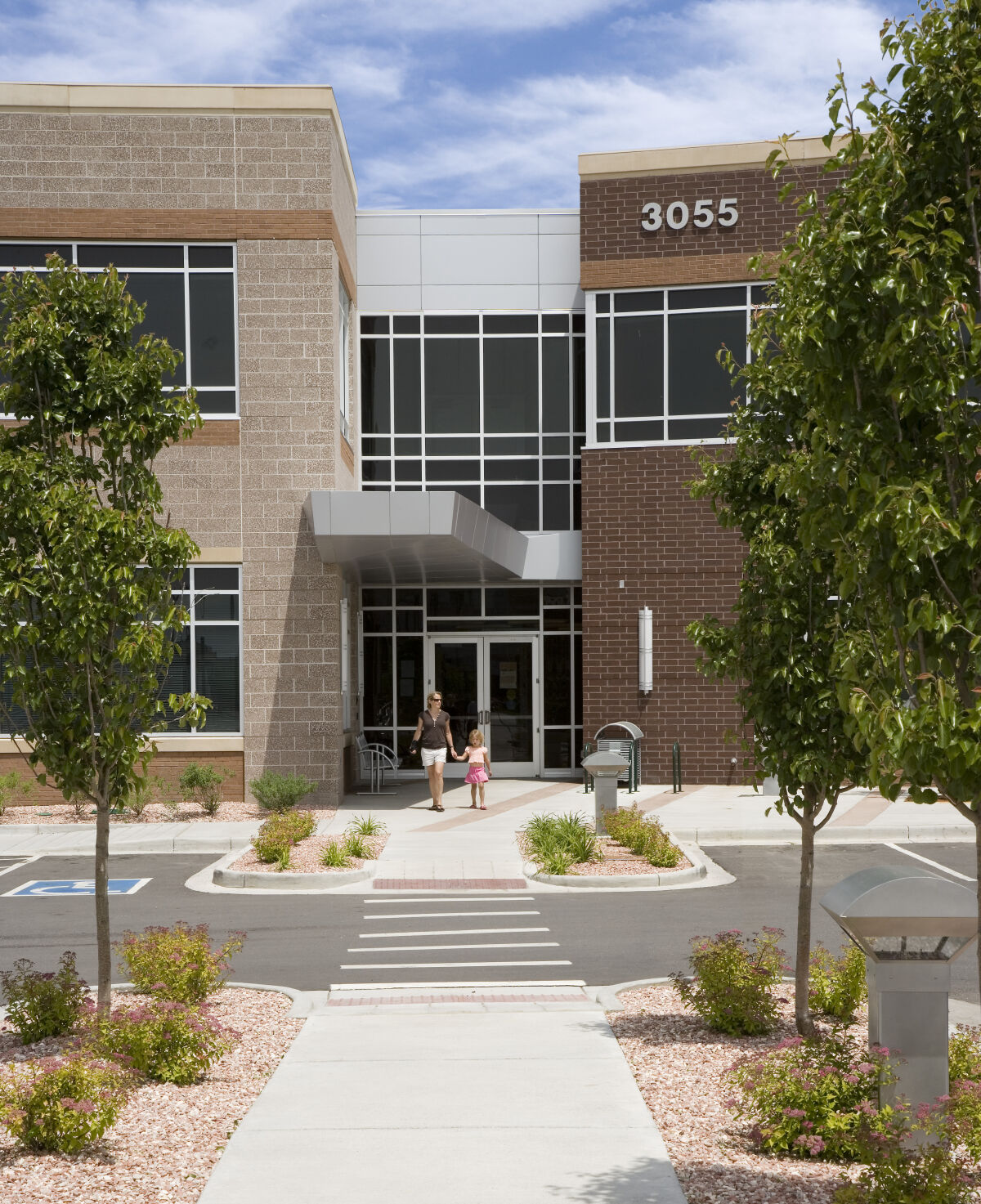 Main entrance of the office building at 3055 Roslyn Street with a concrete walkway, landscaped planting beds, brick and stone façade, and large windows under a partly cloudy sky.