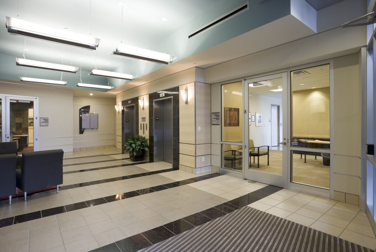 Lobby interior with tiled flooring, wood-accented elevator bays, a seating area with armchairs, overhead linear lighting, and a glass-enclosed conference room.