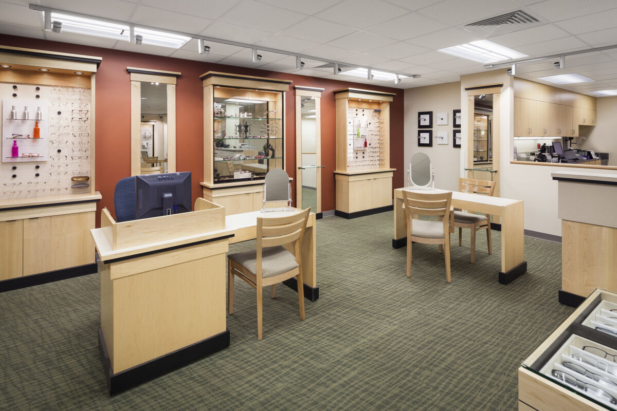 Optical retail area with display cases for eyeglasses, wood cabinetry, framed wall-mounted eyewear displays, consultation tables with chairs, and carpeted flooring under recessed and track lighting.