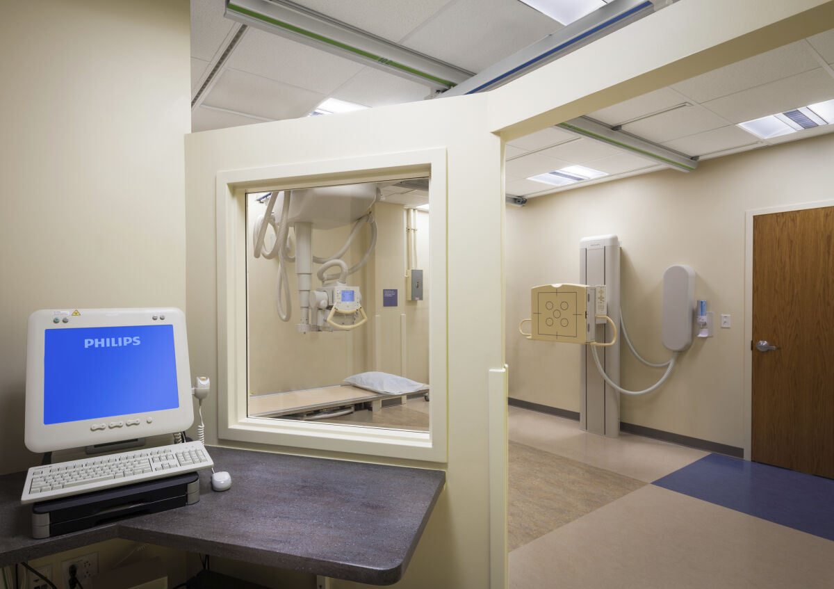Radiology room with an X-ray table and overhead imaging equipment, beige wall panels with mounted components, a wood door, and an adjacent control area with a computer workstation behind a leaded glass window.