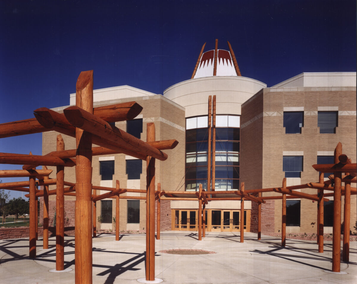 Exterior of the Nighthorse Campbell Native Health Building featuring a central circular tower with patterned crown, large curved glass entry façade, and an outdoor plaza with tall intersecting timber posts.