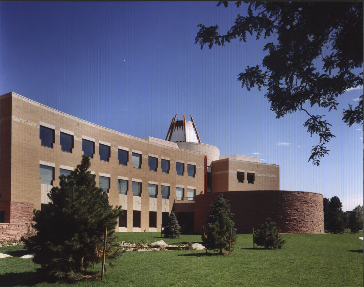 Exterior view of the Nighthorse Campbell Native Health Building featuring a curved brick façade, a round stone-clad pavilion, and a central tower with a patterned crown, set beside landscaped lawns and trees.