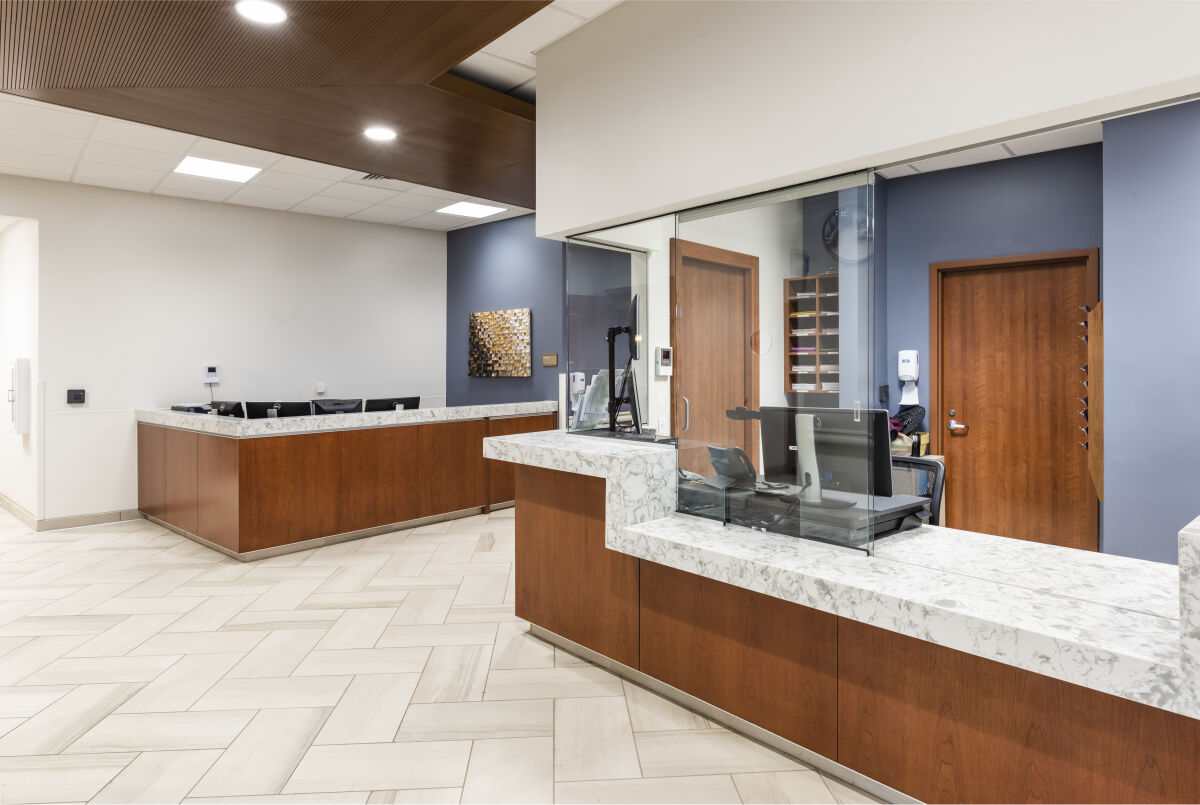 Reception area with two marble-topped wood counters, glass partitions at the registration desk, wood doors leading to clinical rooms, blue accent walls, and herringbone tile flooring under recessed ceiling lighting.