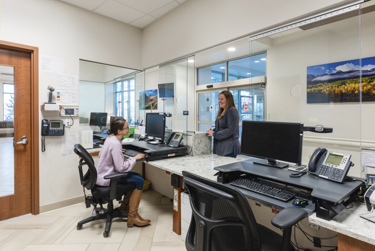 Registration workstation with a marble-topped counter, multiple computer monitors, wall-mounted phones, glass privacy partitions, wood door, and framed landscape artwork along the wall.