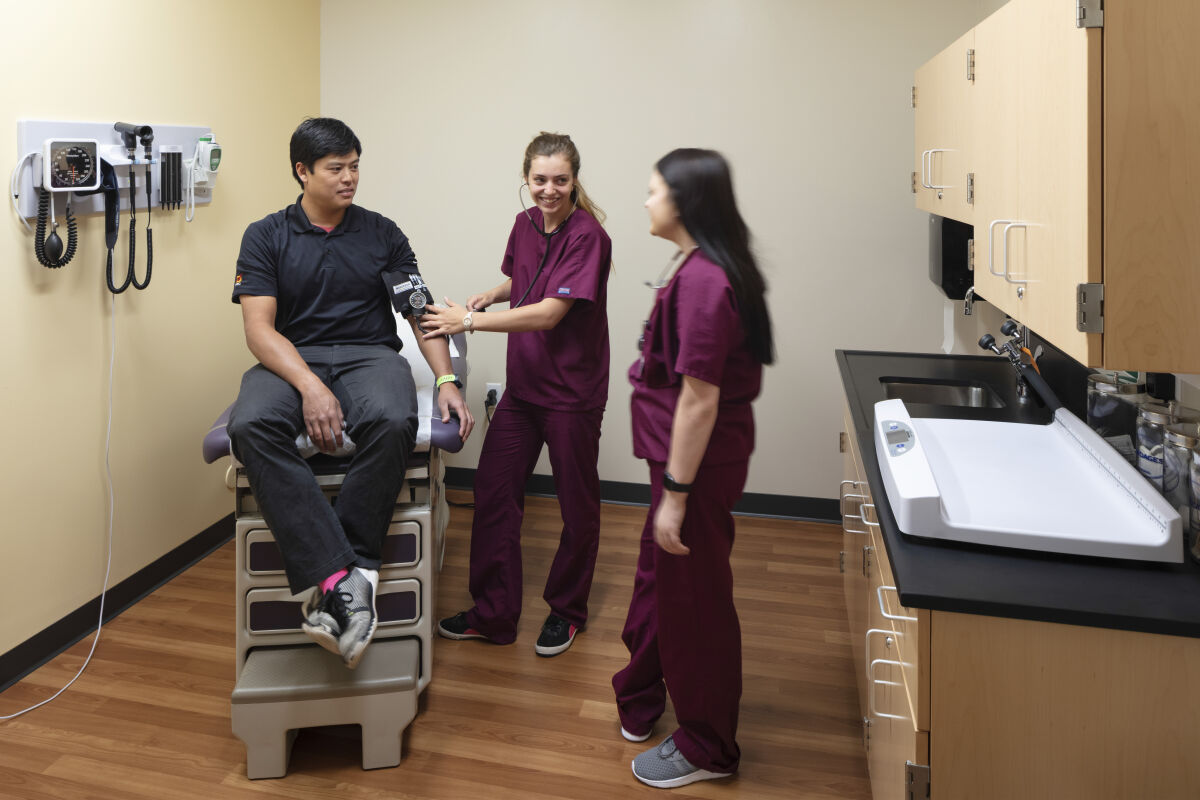 Exam room training lab with an adjustable exam chair, wood cabinetry with a sink and gloves dispenser, wall-mounted diagnostic equipment, polished wood-look flooring, and beige and yellow painted walls.