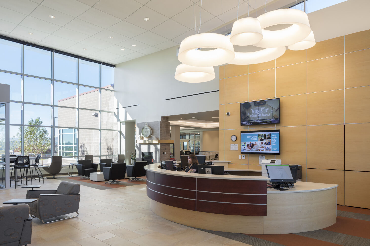 Lobby featuring a curved wood reception desk with computer workstations, large floor-to-ceiling windows, lounge seating areas, tiled flooring, pendant ring light fixtures, and light wood wall panels with mounted digital screens.