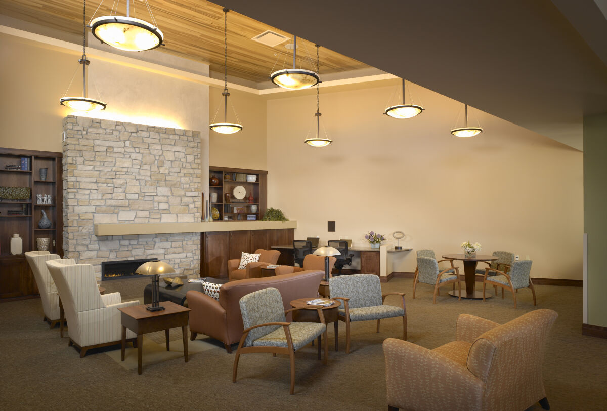 Family lounge with a stone fireplace, wood built-in shelving, upholstered lounge chairs and sofas, round wood tables with chairs, pendant ceiling lights, and carpeted flooring beneath a wood-accent ceiling.