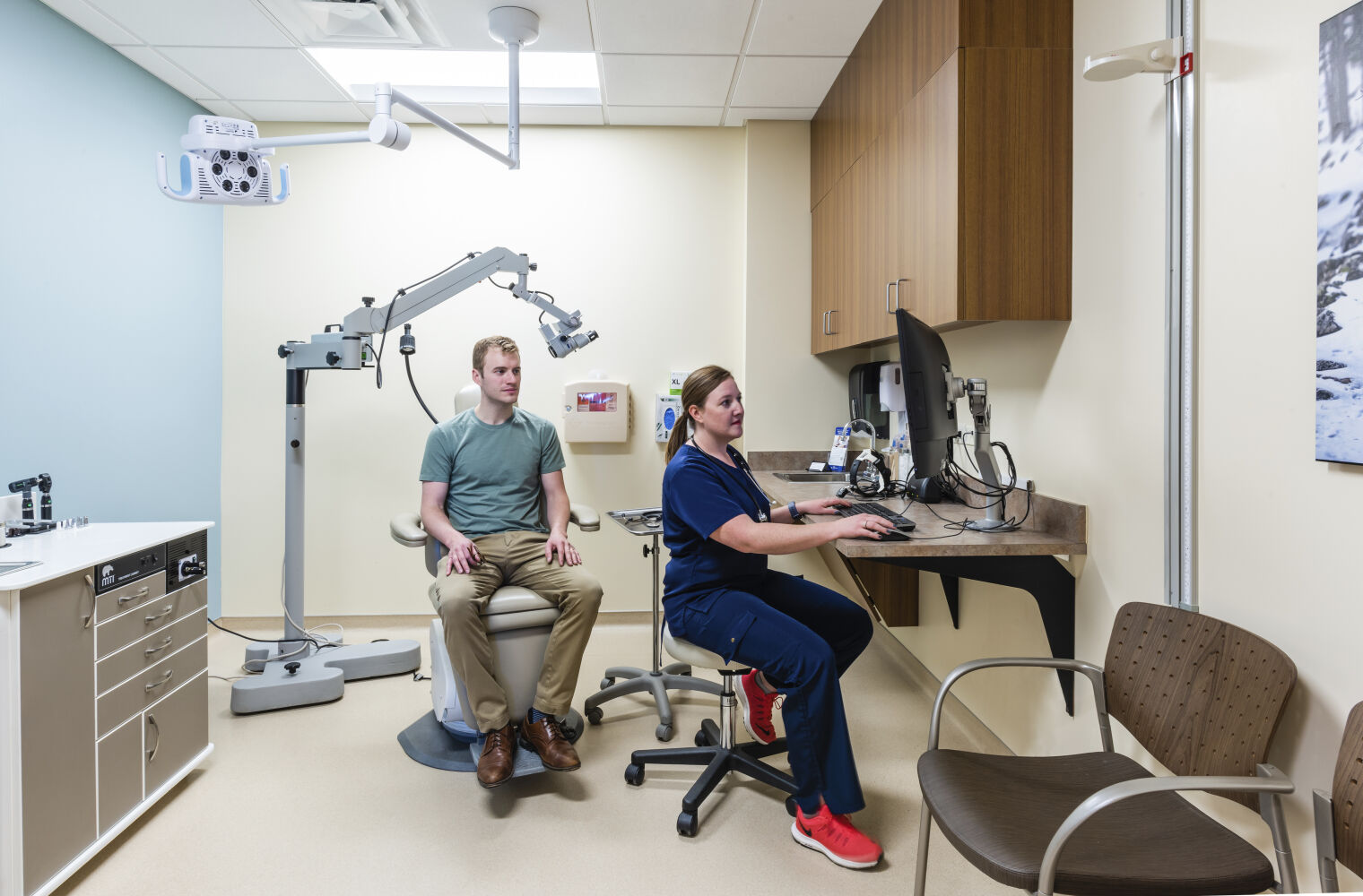 Exam and procedure room with an adjustable patient chair, ceiling-mounted examination light, wall-mounted diagnostic equipment, wood cabinetry with upper storage, a corner computer workstation, and beige flooring.