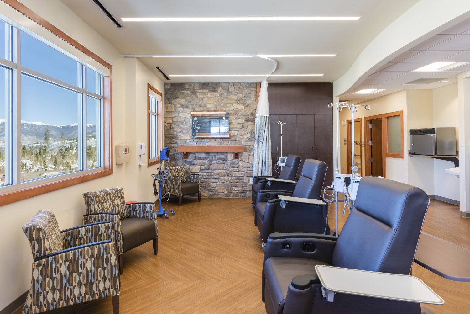Infusion treatment bay with reclining treatment chairs, IV poles, a stone accent wall with a recessed display, wood-look flooring, built-in cabinetry, large windows, and ceiling-mounted lighting.