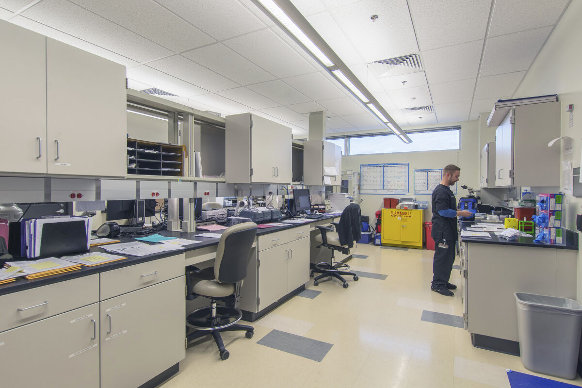 Laboratory interior with workbenches, storage cabinets, computers, analytical equipment, and labeled hazardous‑materials containers.