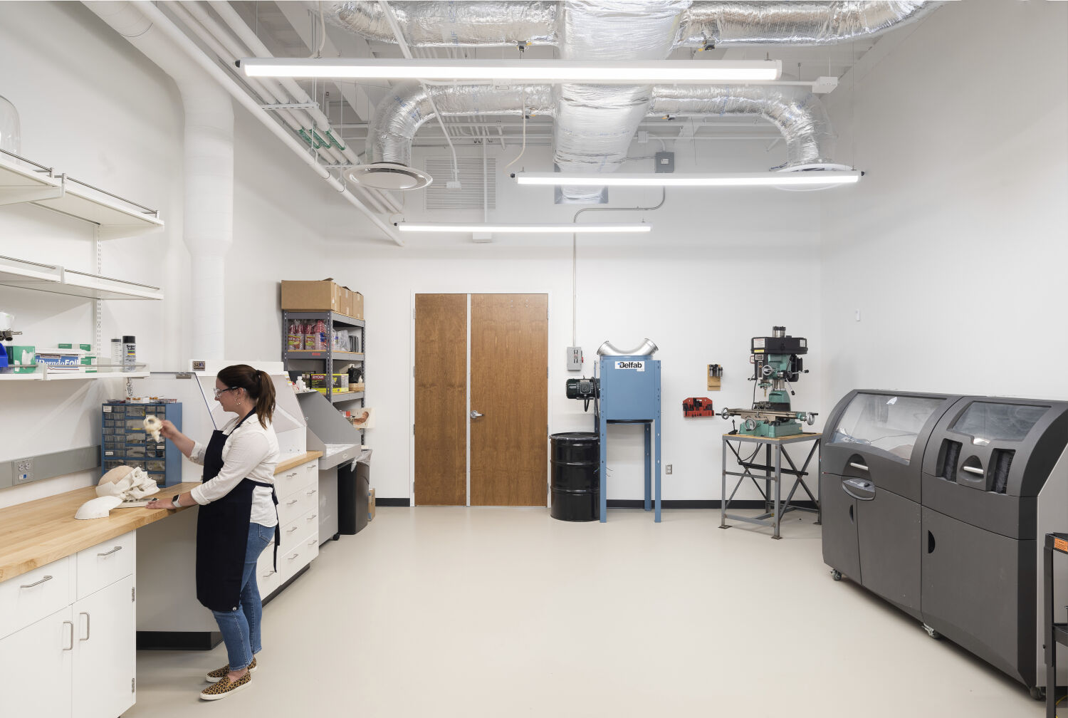 A bioengineering fabrication lab with wood work surfaces, open wall shelving, enclosed machining equipment, large-format fabrication machines, exposed mechanical systems overhead, and wide open floor space for prototyping activities.
