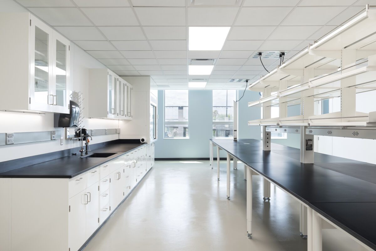 A bioengineering laboratory with long black workbench tables, white perimeter cabinetry with glass‑front uppers, overhead shelving and task lighting, integrated power columns, resilient flooring, and large windows at the end of the room.