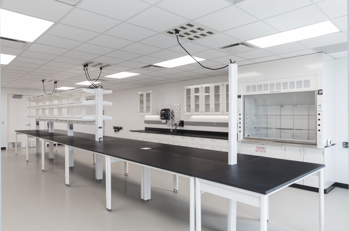 A bioengineering laboratory with long black workbench tables arranged in rows, integrated power columns, overhead shelving, white casework along the perimeter, multiple fume hoods on one wall, and a wide open circulation zone.