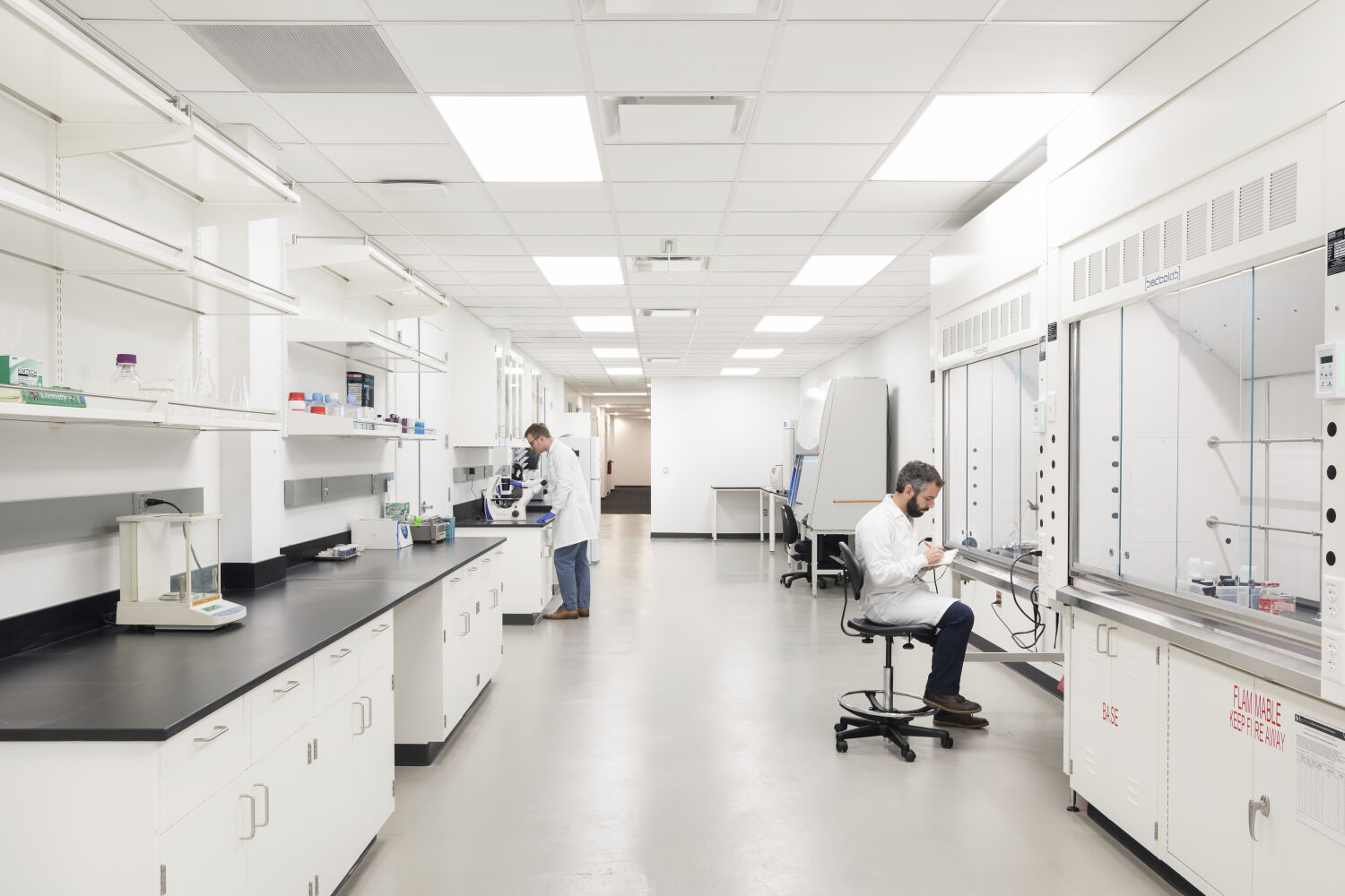 A bioengineering wet laboratory with long bench workstations, black countertops, white casework, multiple fume hoods along one wall, overhead shelving with supplies, and a wide central circulation aisle.