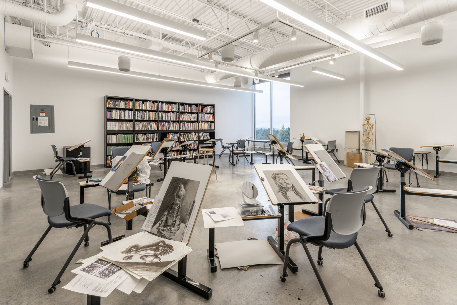 Drawing studio in the Casper College Tobin Visual Arts Center with adjustable drawing desks, rolling chairs, artwork displayed on work surfaces, a large shelving wall filled with books and supplies, polished concrete flooring, and exposed ductwork.