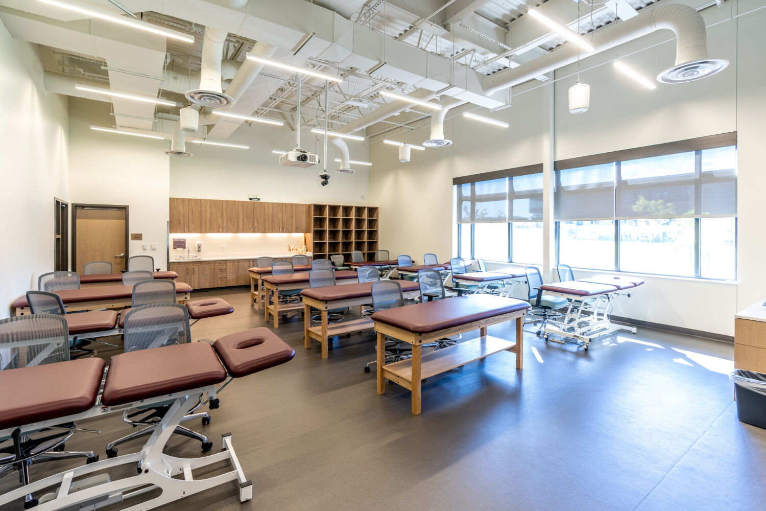 Therapy instruction lab with multiple padded treatment tables, mesh‑back task chairs, wall cabinets with storage cubbies, large windows with roller shades, exposed ductwork, and overhead lighting.
