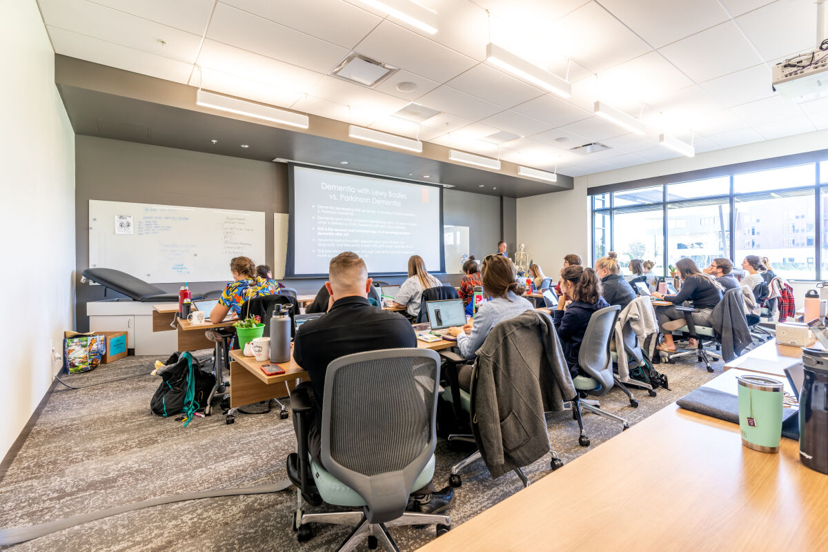 Classroom with rows of tables and task chairs facing a projection screen, a large whiteboard, carpet tile flooring, a ceiling with recessed lighting, and full‑height windows along one side of the room.