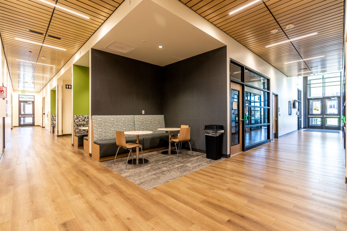 Corridor with a built‑in seating alcove featuring patterned upholstery, small round tables with wood chairs, wood‑pattern flooring, a slatted wood ceiling with linear lighting, and adjacent glass‑fronted rooms.