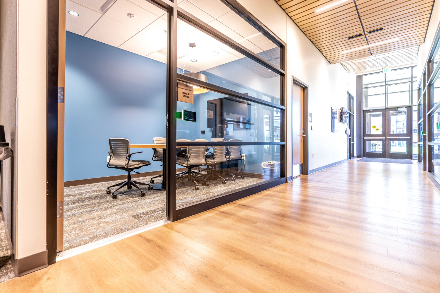 Corridor in the St. Mary’s SCL Health Medical Education Center at Colorado Mesa University with a glass‑fronted group study room containing a wood table, mesh‑back chairs, wall‑mounted display, and overhead lighting.