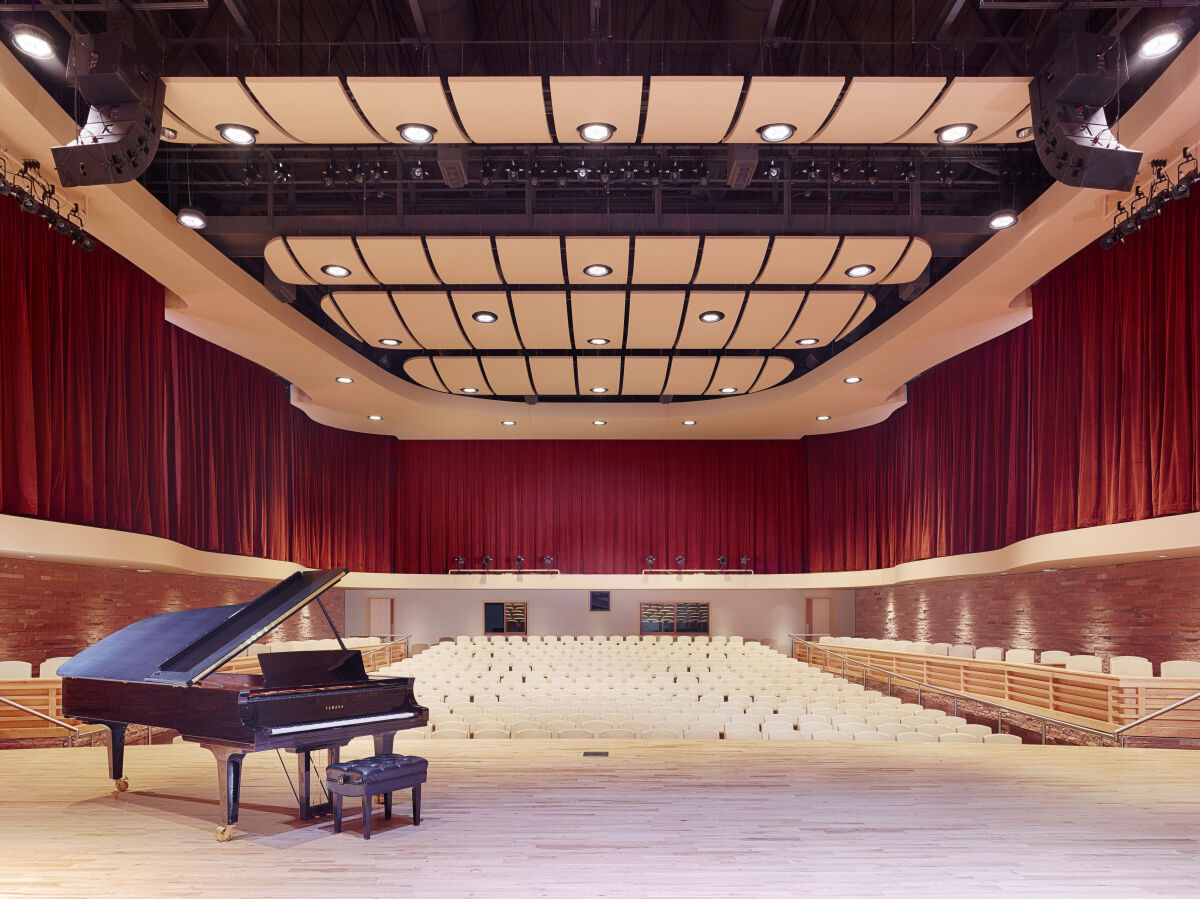 Performance hall with a grand piano on the stage, tiered auditorium seating, red stage curtains surrounding the room, curved acoustic ceiling panels with integrated lighting, and brick accent walls along the lower seating areas.