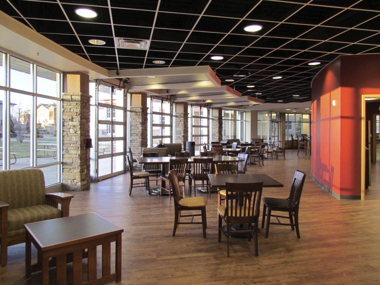 Campus dining and lounge space with wood flooring, round tables, lounge seating, and large windows along the perimeter.