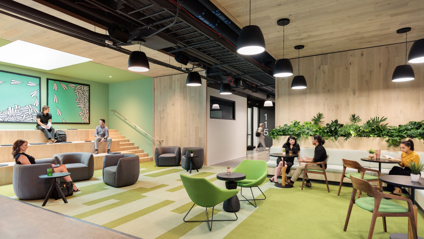 Open commons at Infinite Labs with tiered seating, lounge furniture, café tables, overhead pendants, and green accent flooring.