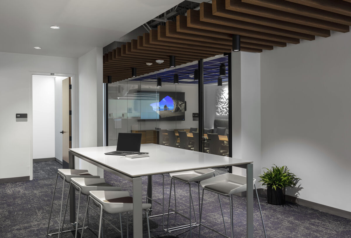 Collaboration area at Cogent Biosciences with a high‑top worktable, wood slat ceiling, glass‑fronted conference rooms, and carpeted flooring.