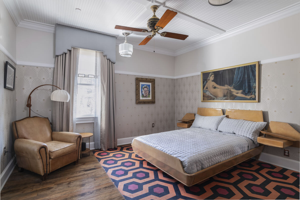 Bedroom in the Stanley Caretaker’s Cottage with a platform bed, patterned wallpaper, hardwood flooring, ceiling fan, and vintage furnishings.