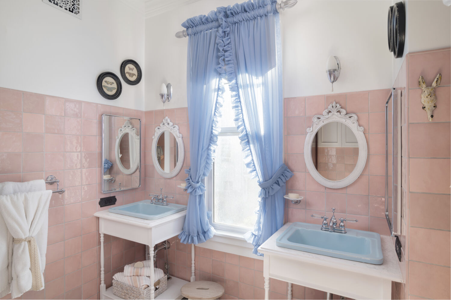 Bathroom in the Stanley Caretaker’s Cottage with pink wall tile, dual pedestal sinks, decorative mirrors, and a window with ruffled curtains.