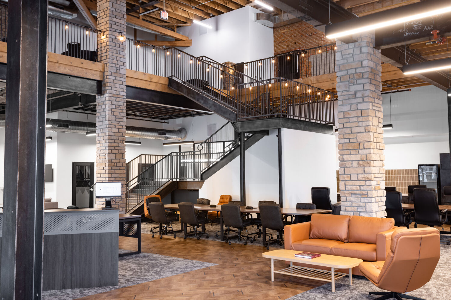 Interior view of the Casper Citiwerks coworking space showing a meeting area with long tables, lounge seating, exposed brick columns, and a central staircase leading to the mezzanine.