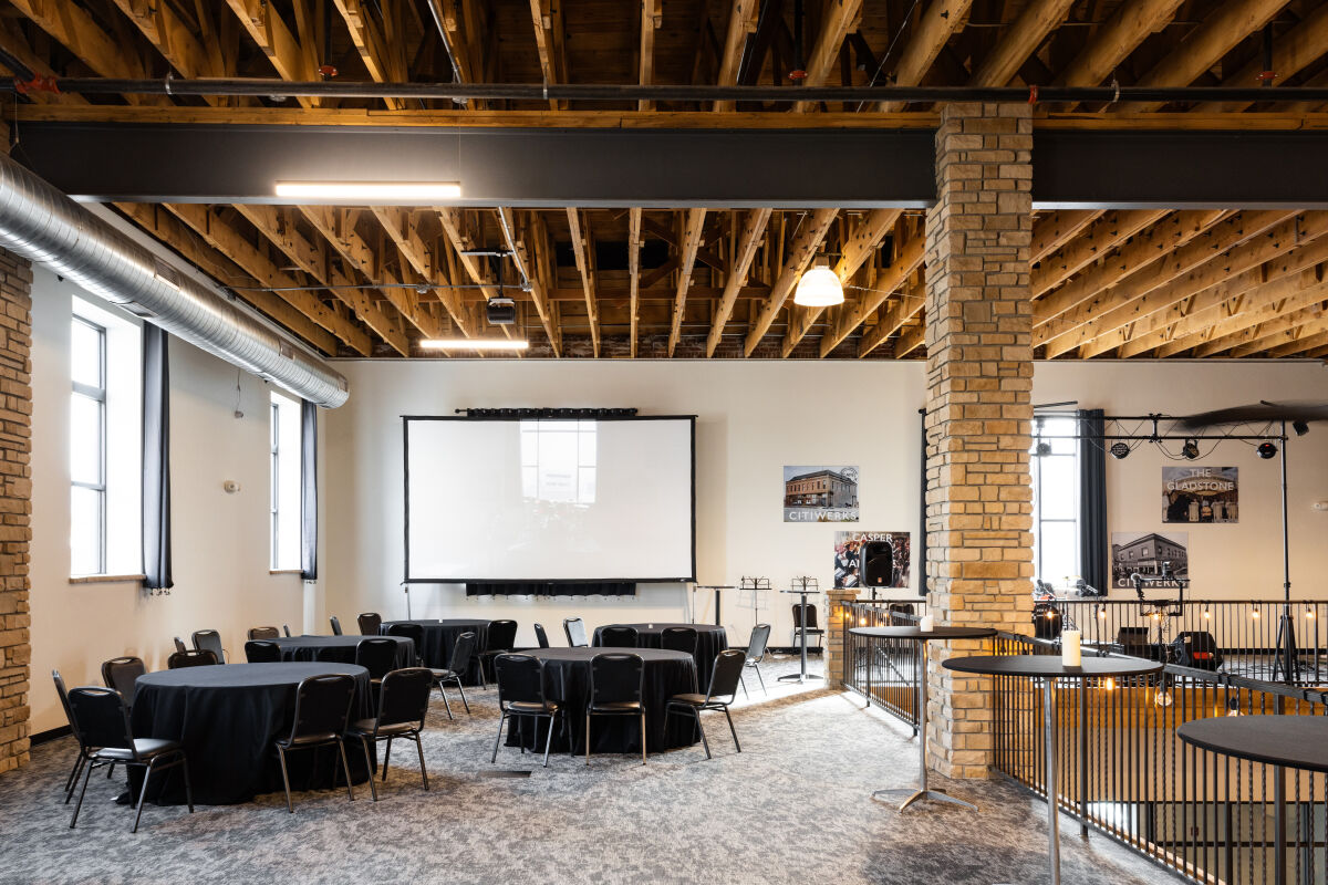 Casper Citiwerks mezzanine event space arranged with round tables and chairs, a projector screen, exposed wood ceiling beams, brick columns, and railing overlooking the coworking floor below.