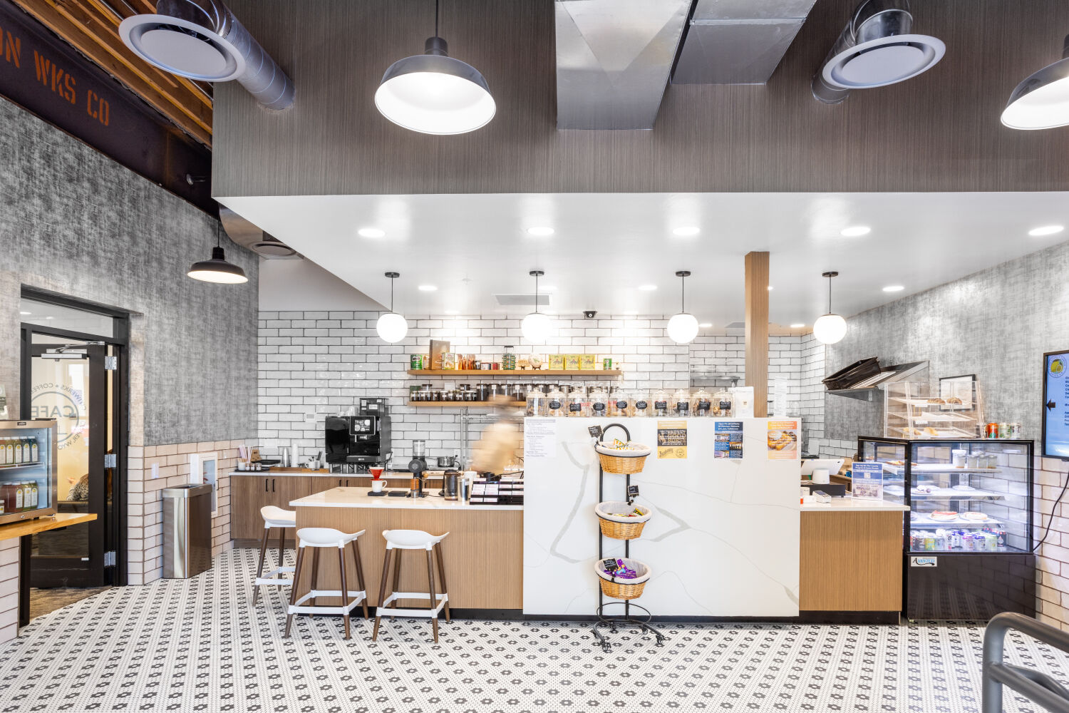 Citiwerks Café with a coffee counter, pastry display case, patterned tile floors, white brick walls, pendant lighting, and counter seating.