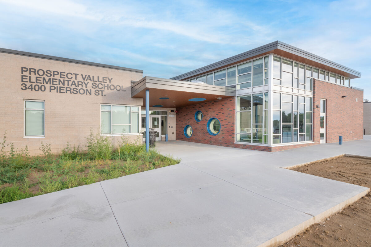 Prospect Valley Elementary School with a recessed main entrance beneath a wood‑clad canopy, brick and light‑colored exterior walls, large glazed windows at the corner, circular accent windows near the entry, and school signage mounted on the facade.