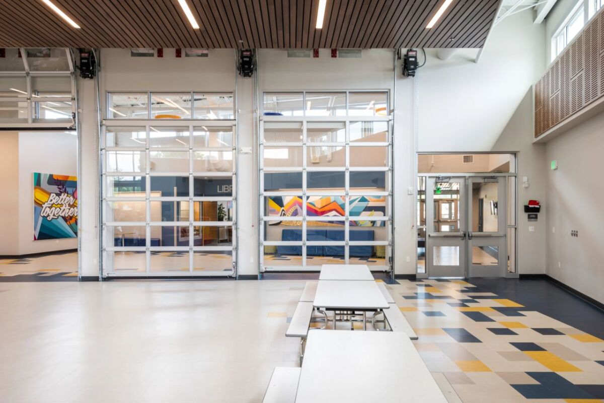 A multipurpose commons space with long cafeteria tables and benches, large glazed overhead doors opening to adjacent learning areas, patterned resilient flooring, wood‑slat ceiling panels, and colorful wall artwork framing the circulation zone.