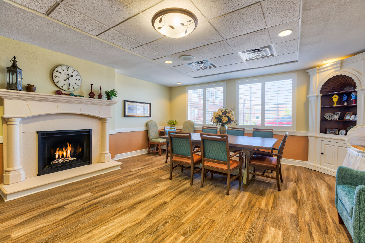 Community dining room at Lincoln Meadows Senior Living with a fireplace, long dining table, upholstered chairs, built‑in shelving, and wood‑look flooring.
