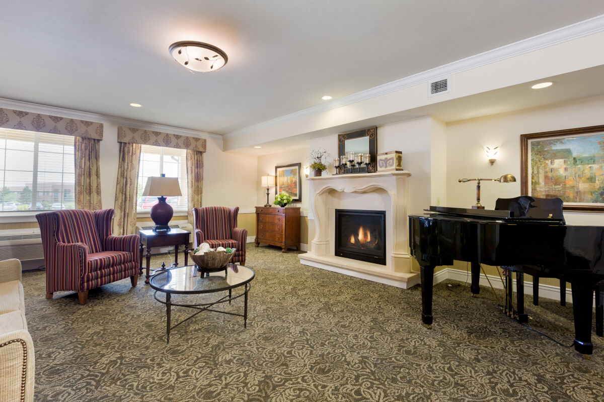 Community lounge at Lincoln Meadows Senior Living with a fireplace, grand piano, upholstered seating, side tables, and patterned carpet.