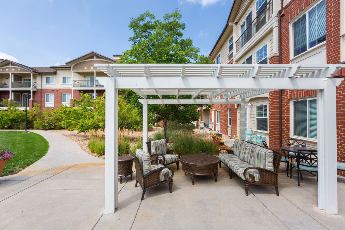 Outdoor seating area at Lincoln Meadows Senior Living with a white pergola, cushioned lounge furniture, paved patio, and surrounding landscaped walkways adjacent to residential buildings.