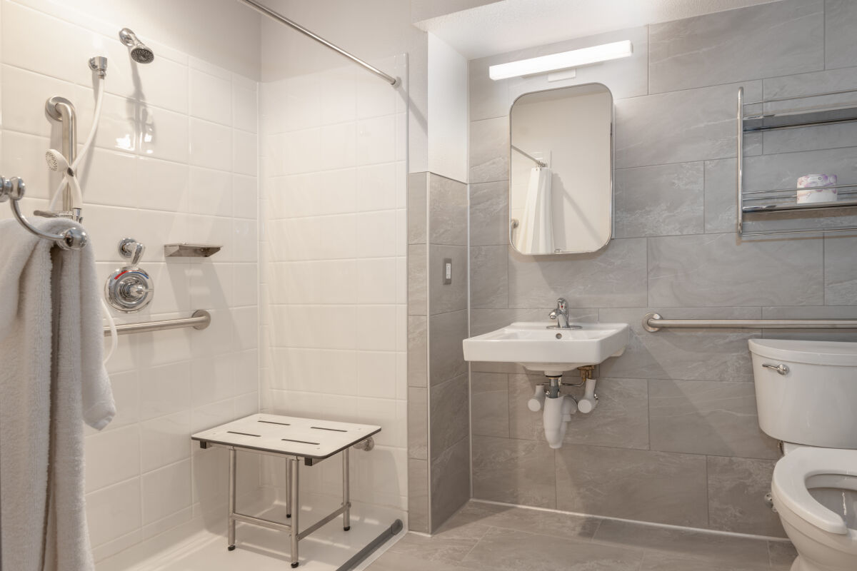 An ADA‑accessible restroom with a roll‑in shower, fold‑down shower seat, grab bars, wall‑mounted sink, toilet with support rails, and slip‑resistant flooring.