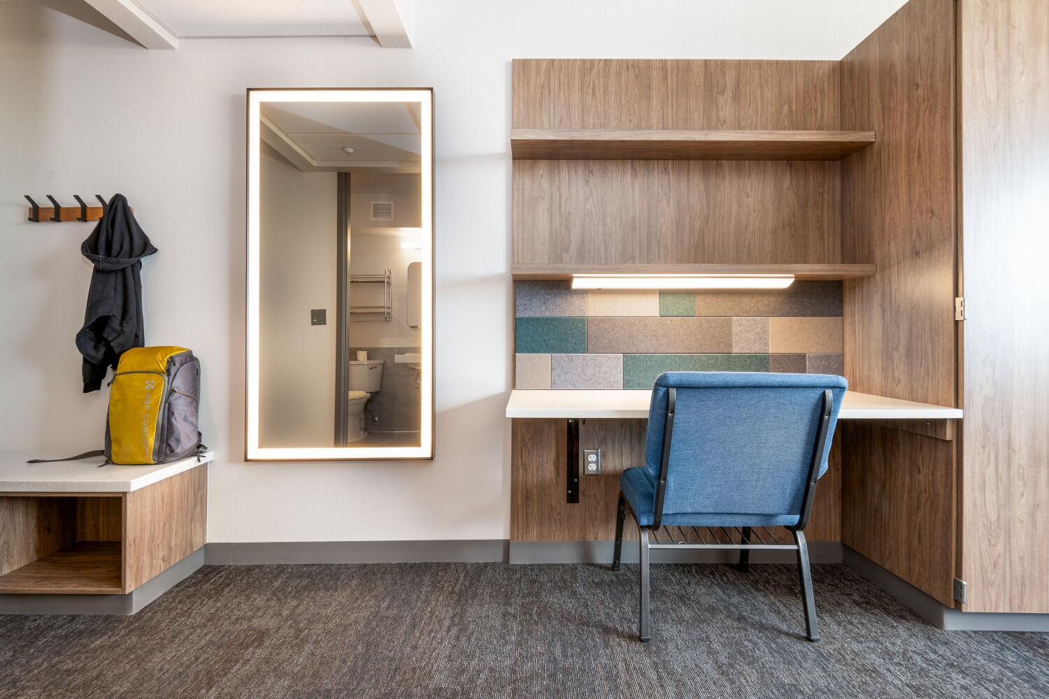 A dorm room entry area with a built‑in bench, open cubby storage, wall hooks, a full‑length mirror, and adjacent shelving and desk workspace.