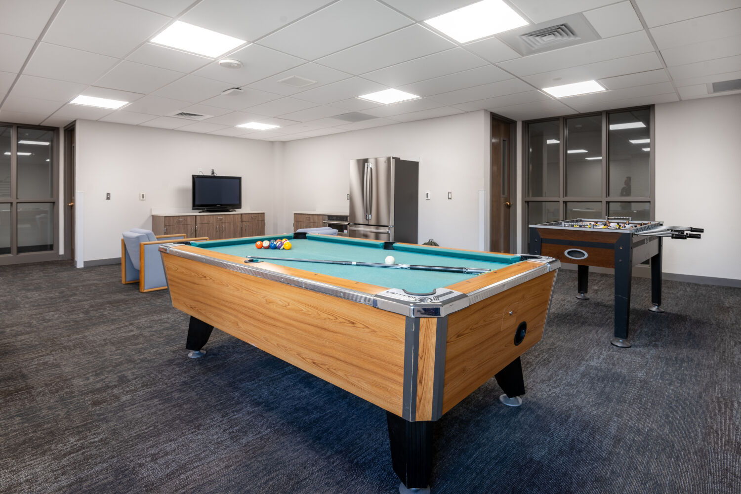 A game room with a pool table, foosball table, seating along the perimeter, storage cabinetry, and a refrigerator arranged within a carpeted space.