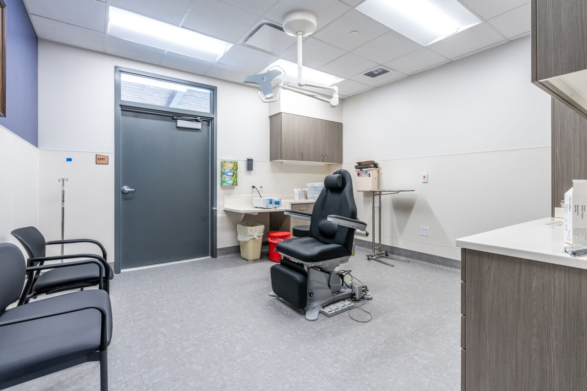 A clinical procedure room with an adjustable exam chair, ceiling‑mounted exam light, built‑in cabinetry, medical equipment storage, and resilient flooring arranged around a central work area.